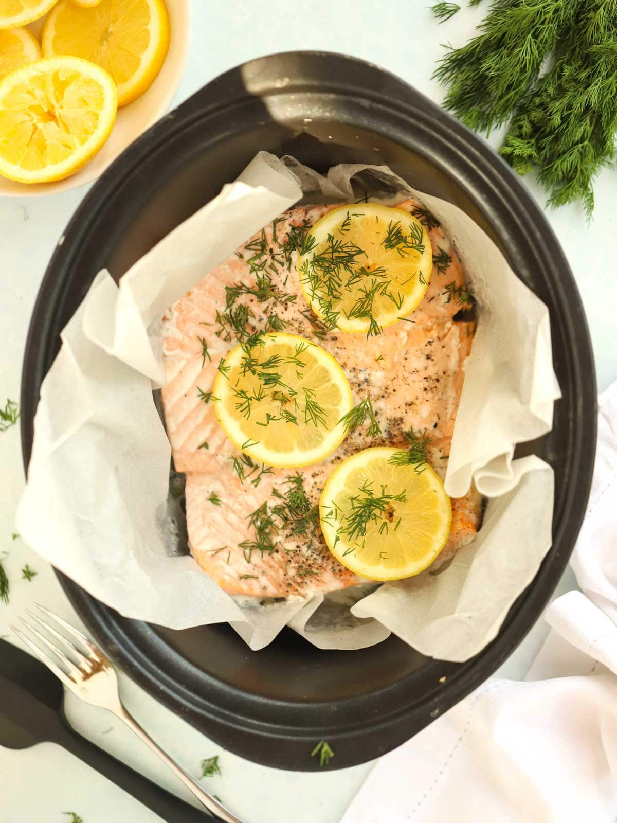 A slow cooker on a white tablecloth with cooked salmon inside, with lemon on top.