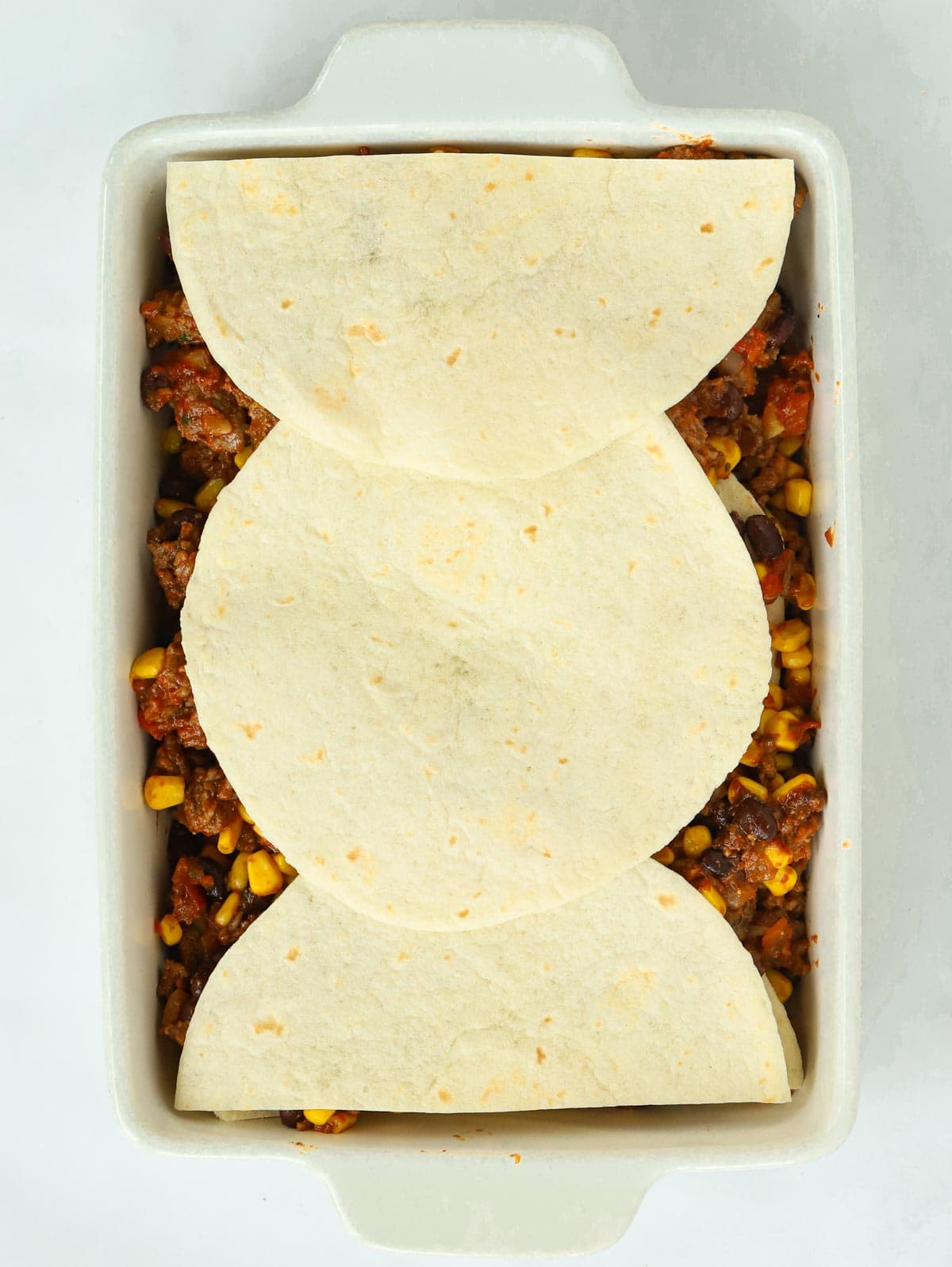 Mexican lasagne with beef mixture topped with uncooked tortillas in a white dish.