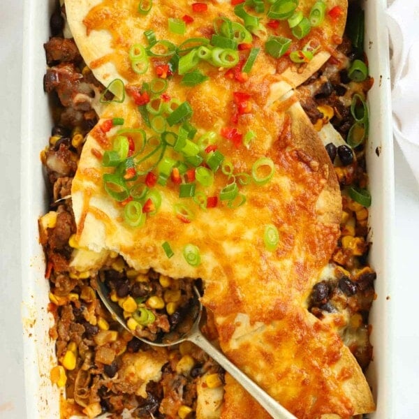Mexican lasagne in a white dish topped with cheese, spring onions and chilli with a spoon lying in the dish.