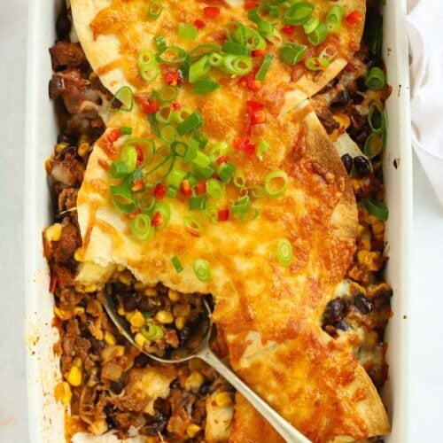 Mexican lasagne in a white dish topped with cheese, spring onions and chilli with a spoon lying in the dish.