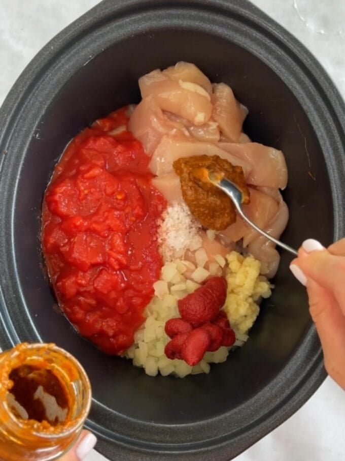 Step 1 of the recipe for slow cooker chicken tikka masala is shown, with the raw ingredients of chicken, tomatoes, onion, tomato puree, salt, garlic and tikka pasta in the pot.