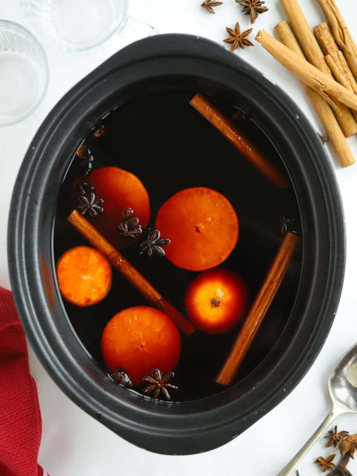 A slow cooker filled with ingredients to make mulled wine, including red wine, oranges and cinnamon sticks.