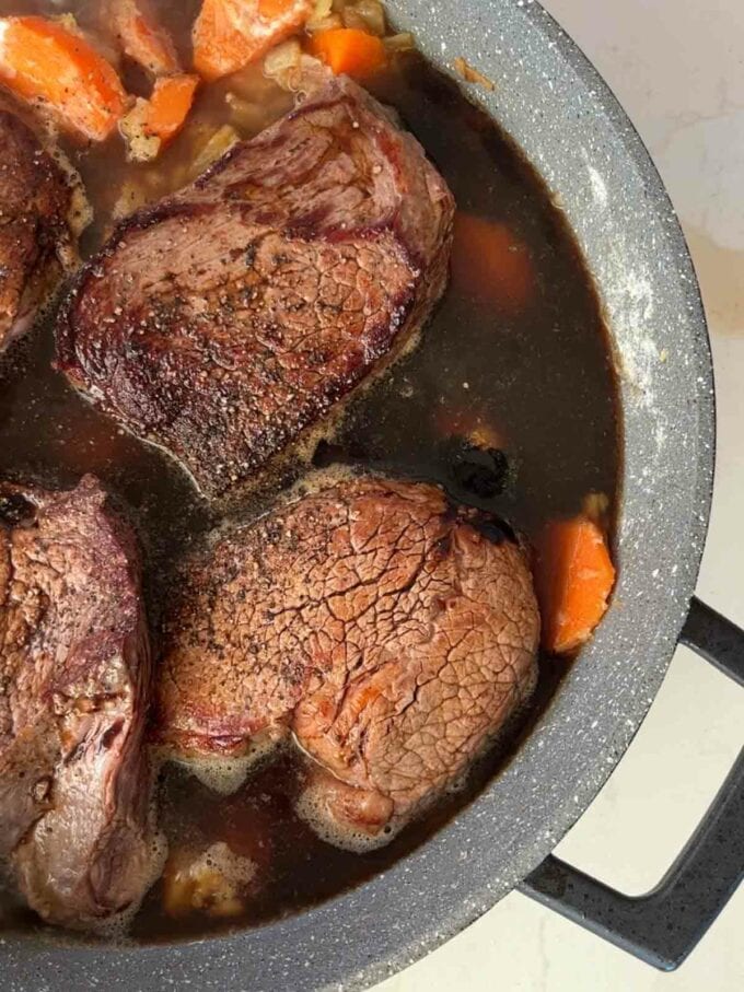 Braised steak cooking in a pan with carrots and onion for the recipe Tender Braised Steak. Step 4.