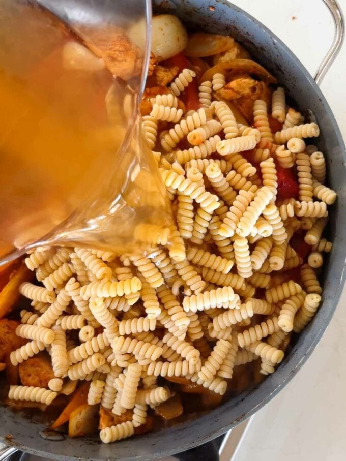 Pasta in a pan with ingredients and stock being poured over for a chicken fajita pasta bake recipe.