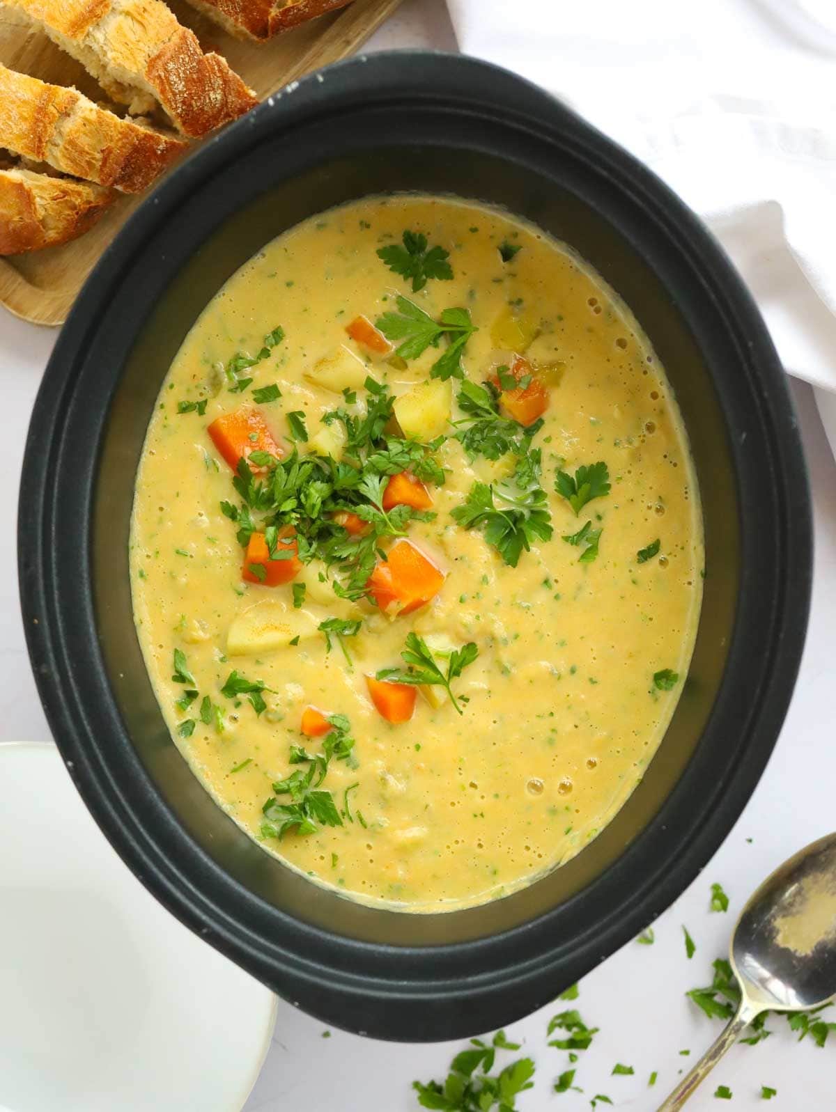 A slow cooker pan with cooked vegetable soup on the table, served with crusty bread.