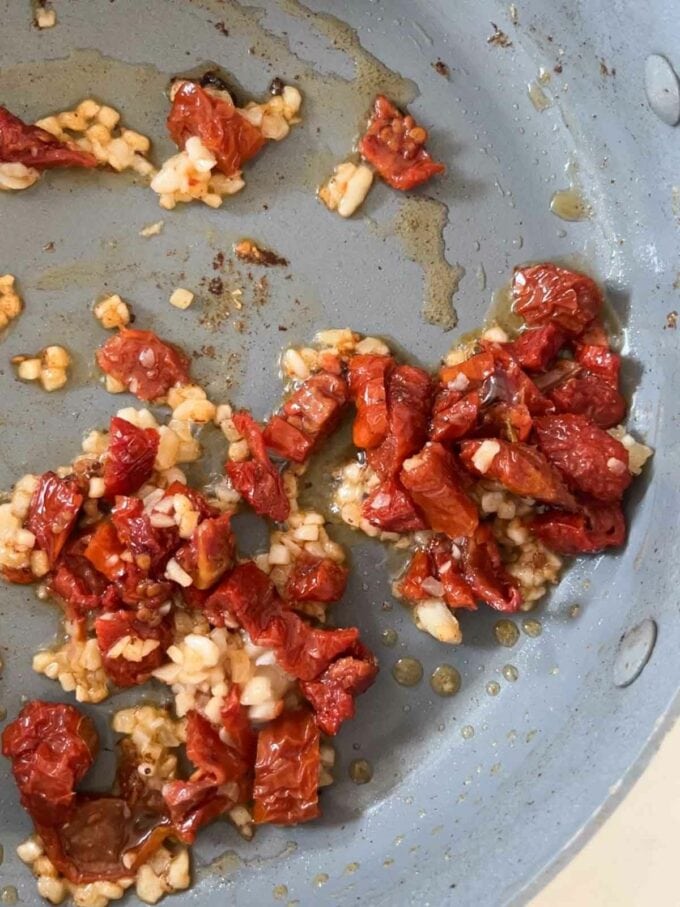 Garlic and chopped sun dried tomatoes being cooked in oil in a pan for the recipe Marry Me Chicken. Step 3.