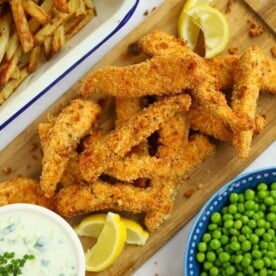 Homemade salmon fish fingers on a serving board, with a bowl of peas, tomato ketchup, tar tar sauce and homemade chips on the side.