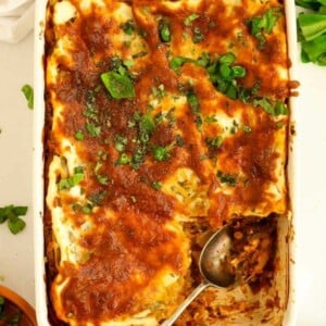 A lasagne dish filled with cooked Courgette Lasagne, topped with cheese and basil. One corner is missing, having already been served with a spoon.
