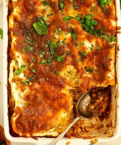 A lasagne dish filled with cooked Courgette Lasagne, topped with cheese and basil. One corner is missing, having already been served with a spoon.