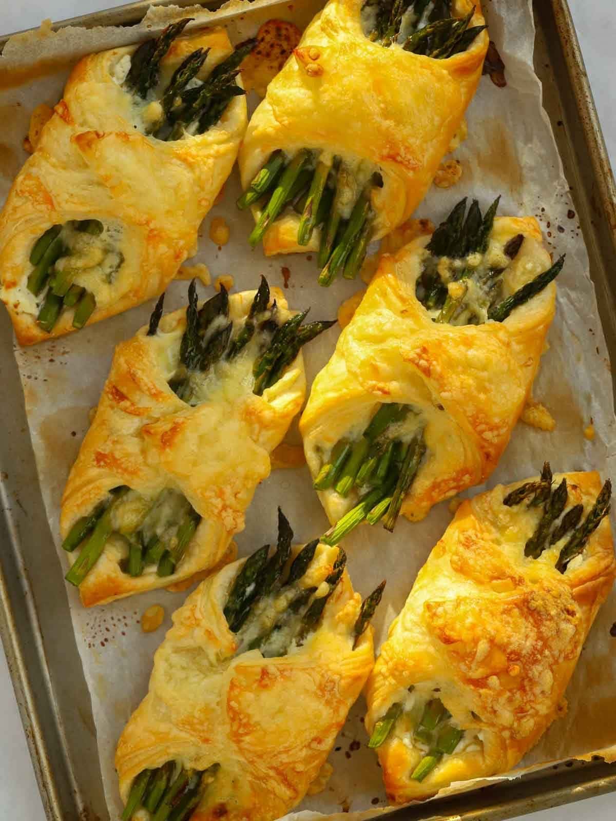 Puff pastry nibbles with asparagus and cheese inside on a baking tray, golden brown from the oven.