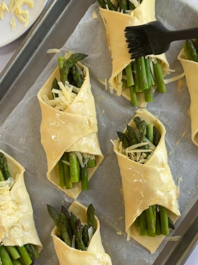 Puff pastry parcels with asparagus and cheese being brushed with egg wash before baking.