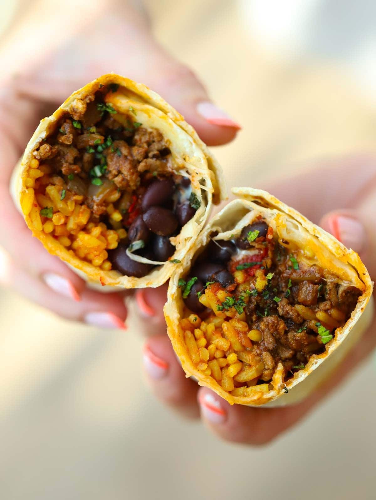 Two hands holding separate halves of a Beef Burritos, filled with mince, rice, beans and cheese.