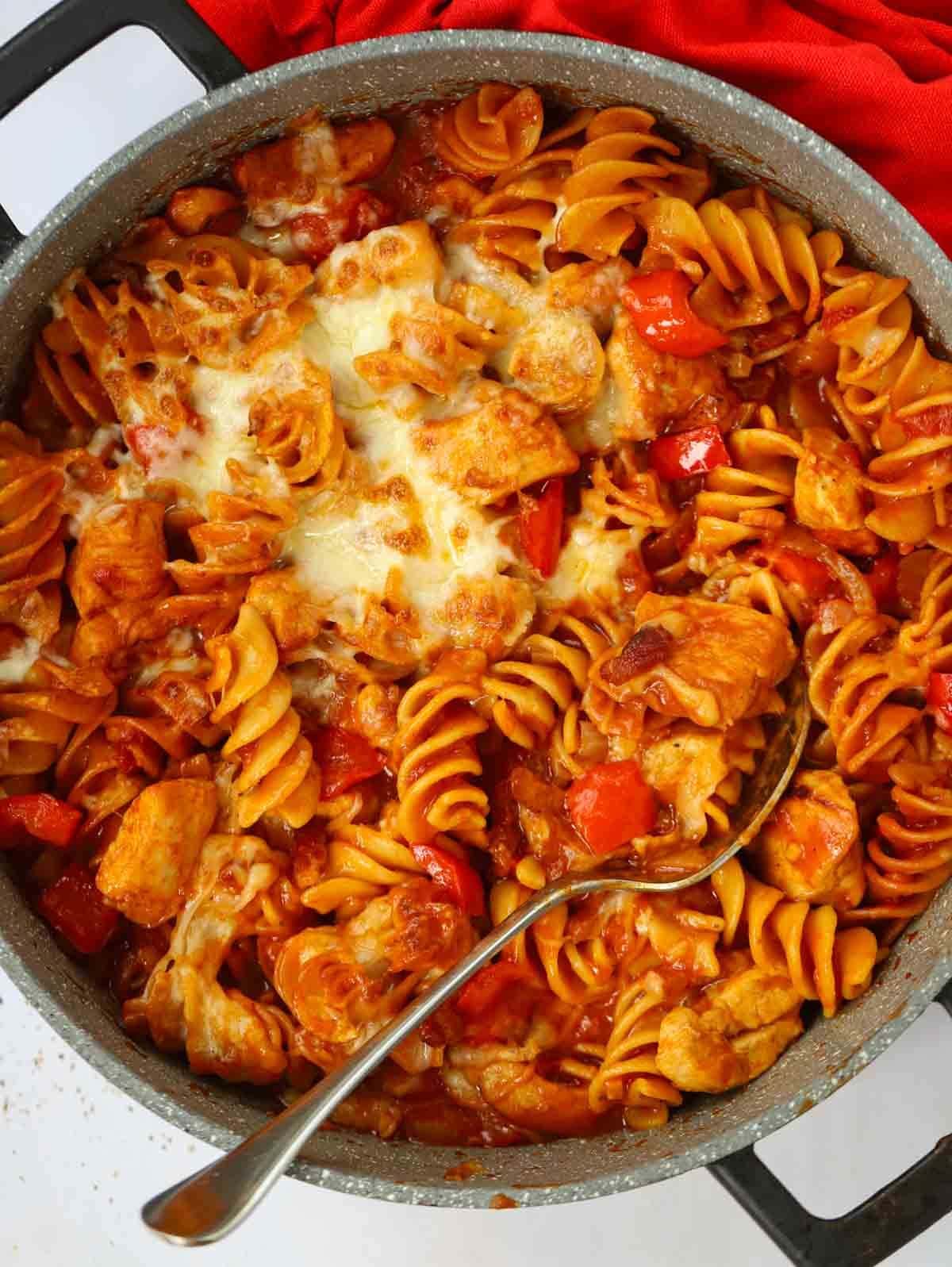 A big pan filled with cooked barbecue chicken and bacon pasta, with a spoon dipped in ready to serve.