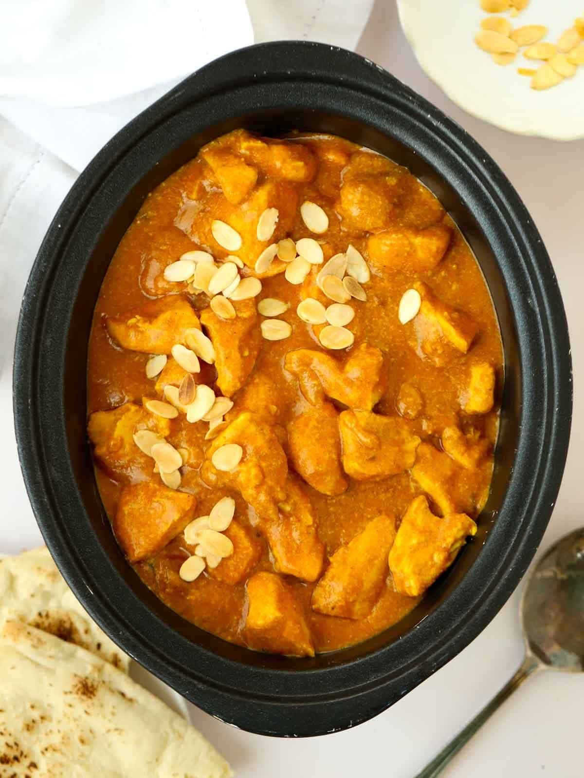 A slow cooker pot filled with cooked chicken korma, topped with almonds flakes and with naan breads on the side.