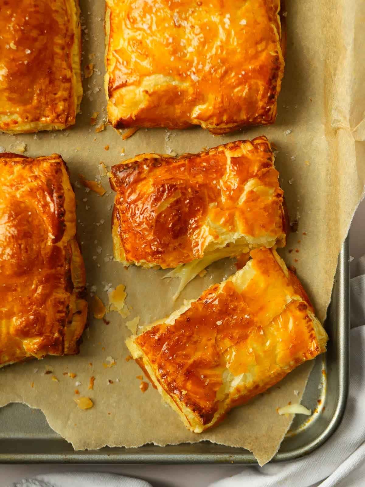 Homemade cheese and onion pasties on a baking tray, one cut in half to show the cheesy filling.