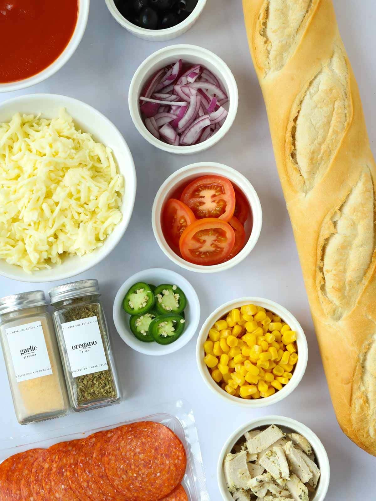 The ingredients for a French Bread Pizza laid on a counter top. Baguette, cheese, herbs and a selection of toppings.