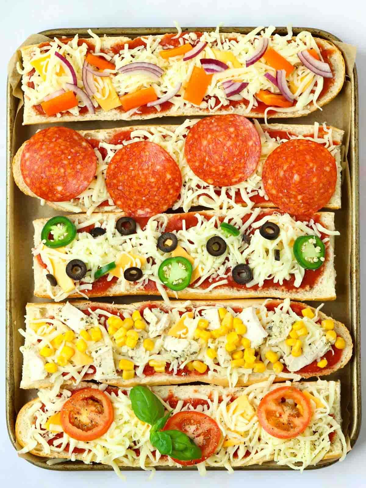Five uncooked French Bread Pizzas on a baking tray with various toppings, ready to go in the oven.