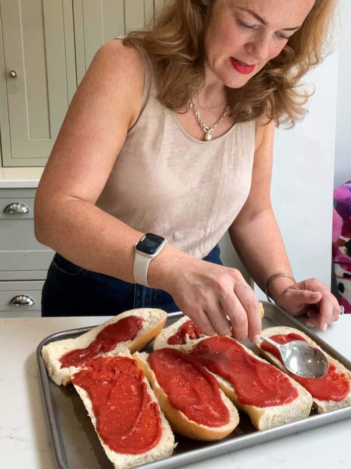 Sarah Rossi of Taming Twins demonstrating how to make french bread pizza step 3. Spreading the tomato base on top of the baguettes.