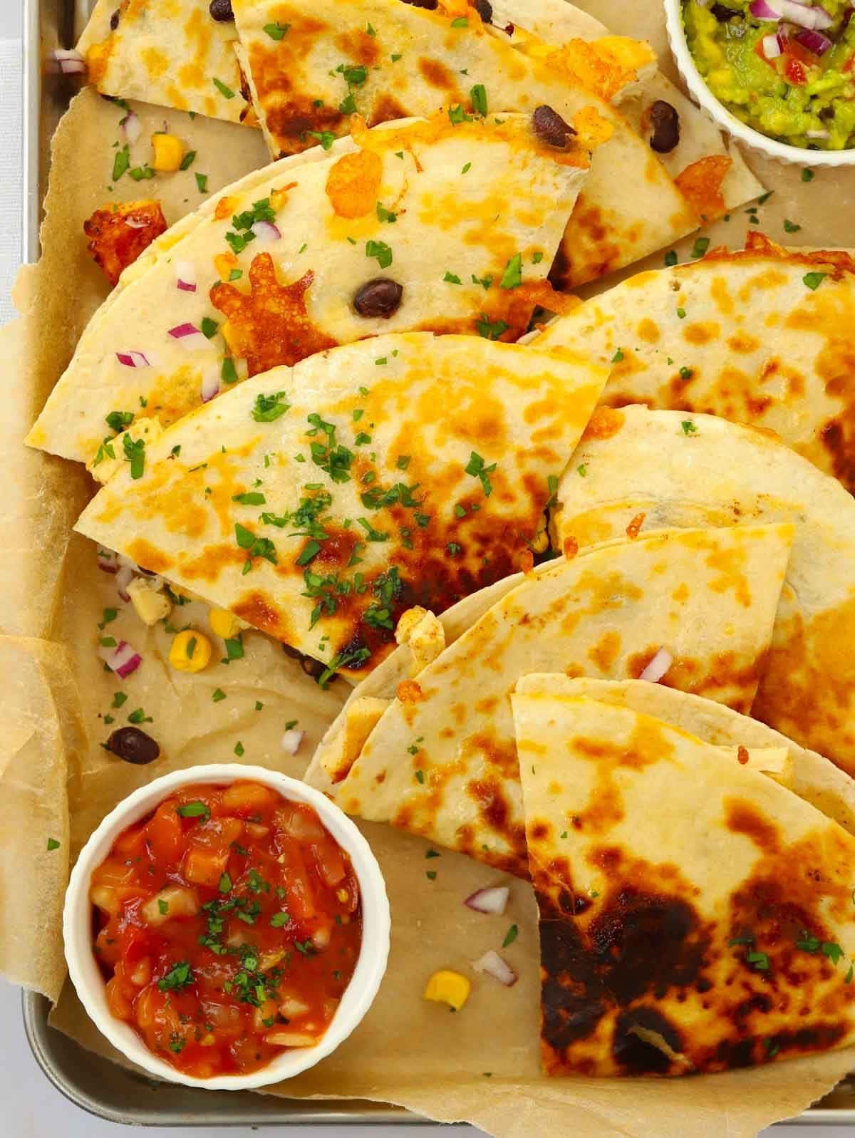 Quartered chicken quesadillas cooked and ready to eat from a baking tray, with sides of salsa and guacamole.