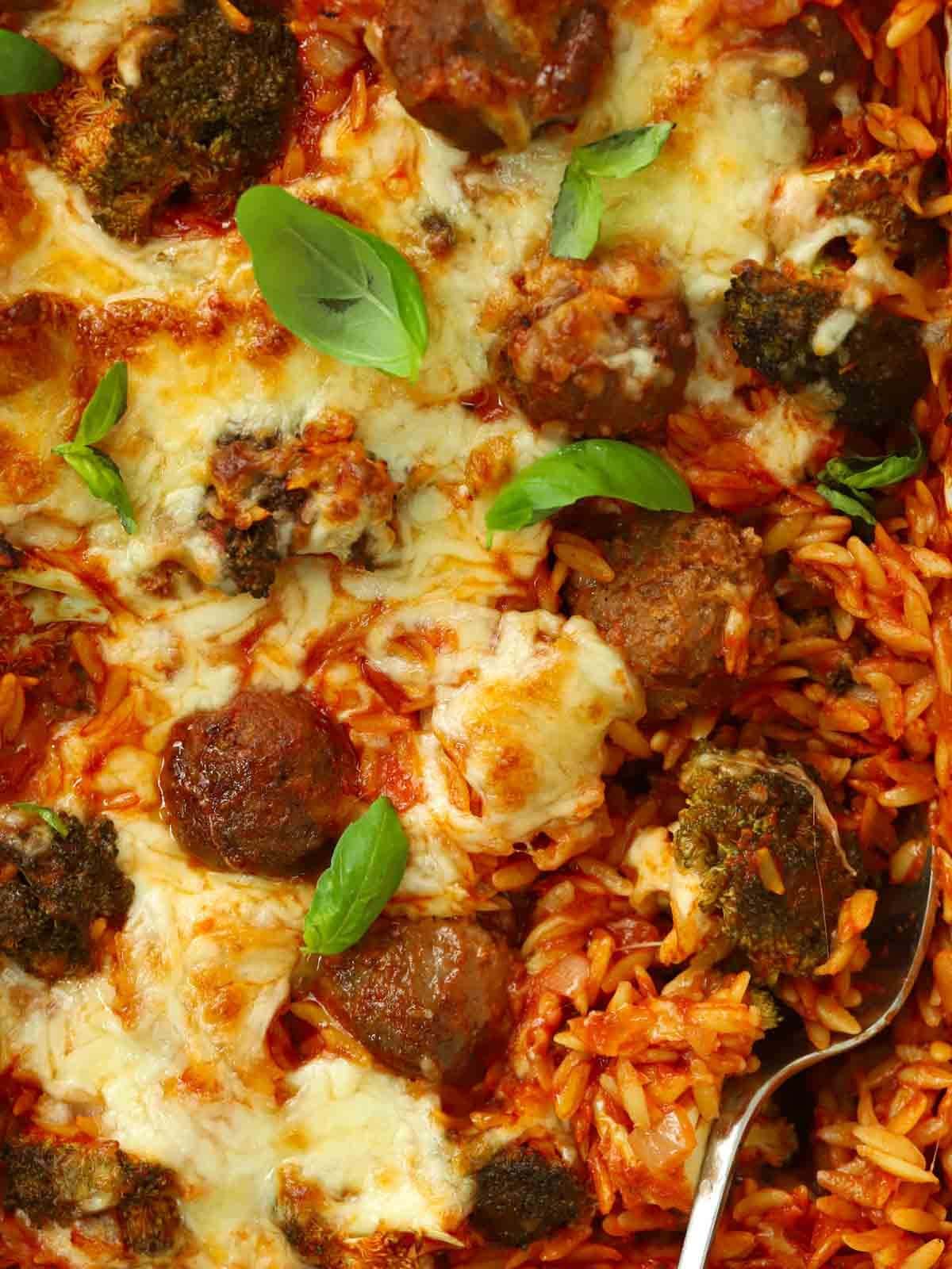 A close up of meatballs, orzo and cheese, cooked in one dish together in the oven. A spoon is showing, ready to serve.