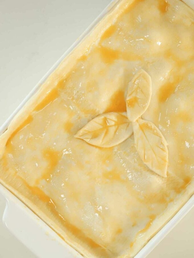 An unbaked chicken pie in a white oven dish, with egg-washed pastry and a leaf cut design, ready to go in the oven.