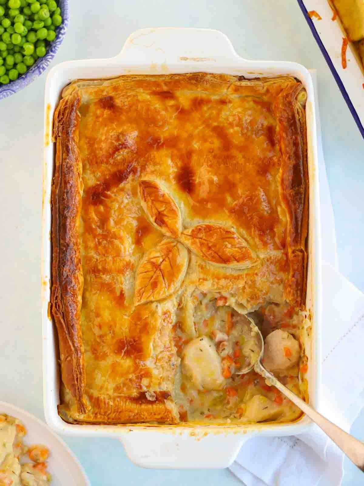 A cooked chicken pie with a corner portion missing, with a spoon ready to serve more. Laid on a white table cloth with a side of peas in a bowl.