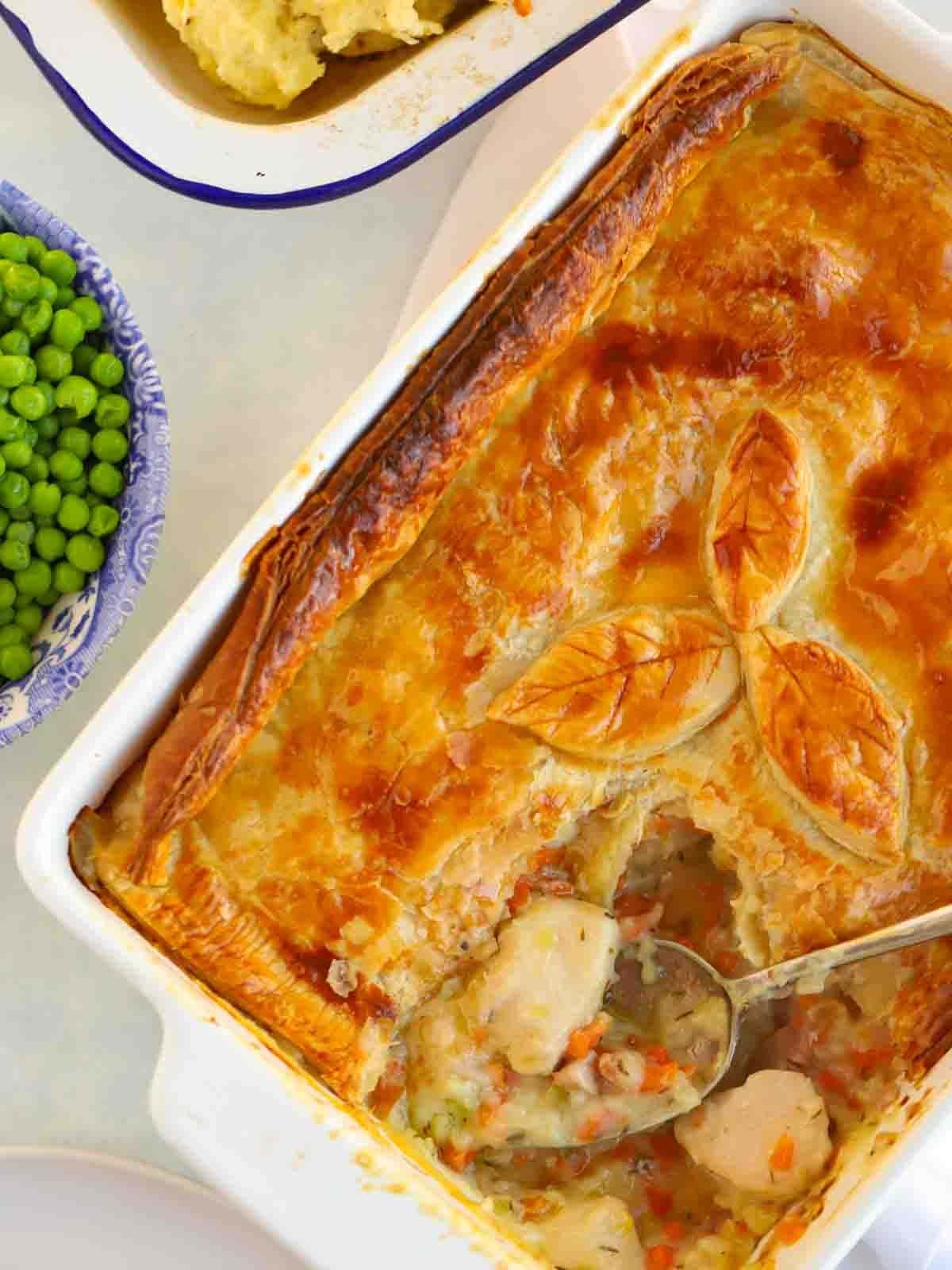 A golden-topped Chicken Pie with a portion missing, on a table with a bow of peas next to it.