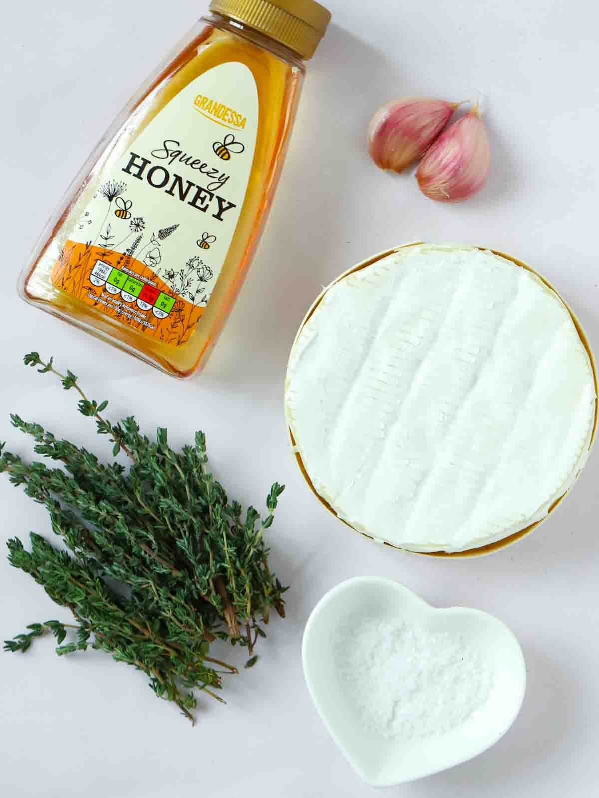 Ingredients for baked camembert laid on a counter. A bottle of honey, uncooked camembert. salt, garlic and thyme.