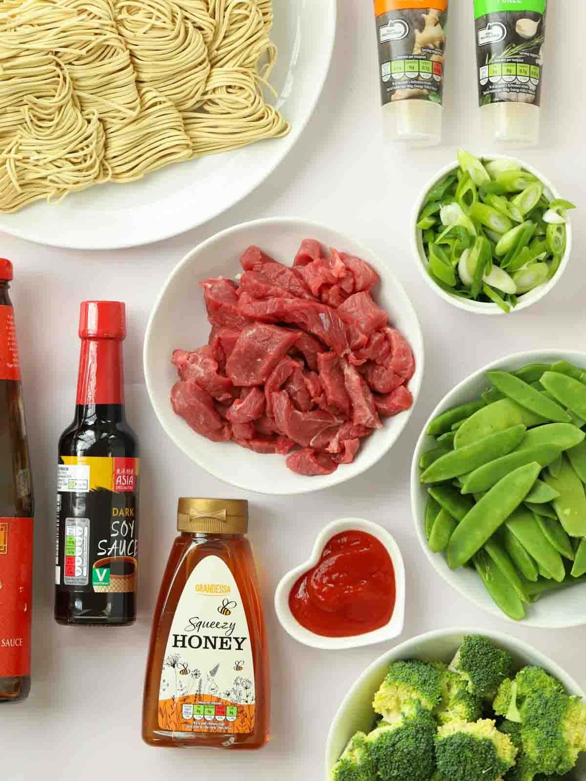 The ingredients for the recipe Sticky Beef & Noodles laid out on a counter top, including steak, green vegetables, sauces and dried noodles.