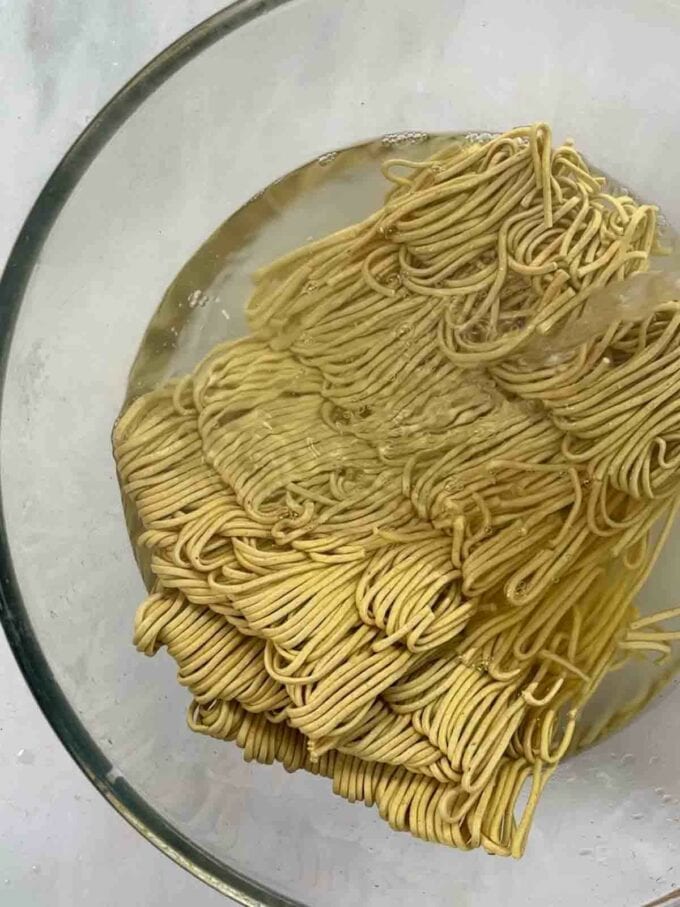 Dried egg noodles soaking in a glass bowl of water.