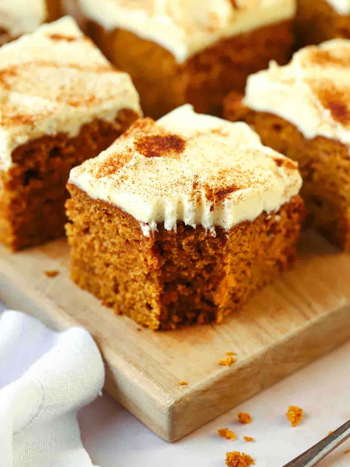 A small bit taken out of the corner of a square of pumpkin cake with cream cheese frosting on top. Other squares in the background on a board.