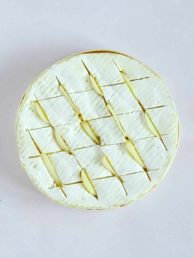 An uncooked round of camembert cheese with slices of garlic pressed into the scored criss cross lines in the rind.