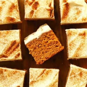 Nine squares of pumpkin cake on a board, with the middle one on its side to show the inside. All of them are topped with cream cheese frosting.