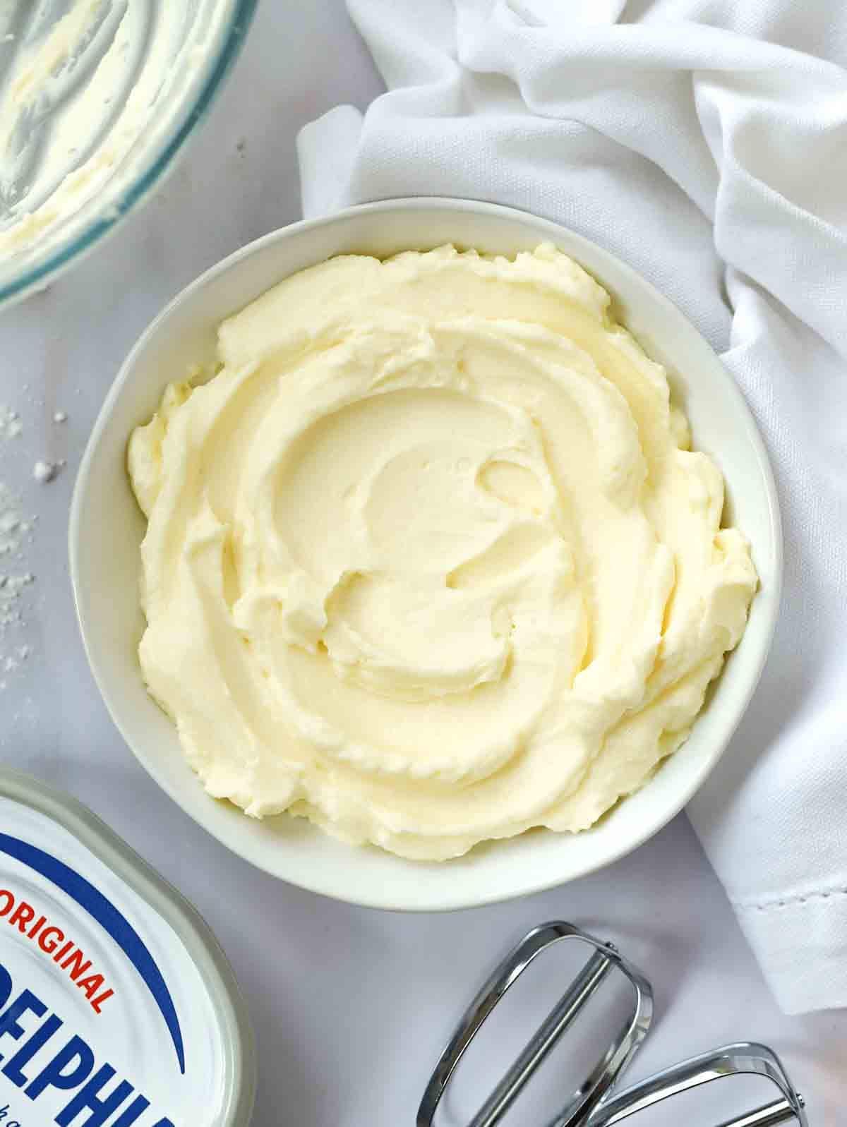 A bowl on a white table cloth, filled with cream cheese frosting, surrounded by a bowl and pack of cream cheese and a whisk.