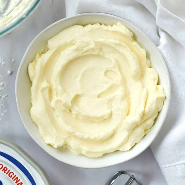 A bowl of cream cheese icing in a bowl on a white table cloth. Just out of sight are the ingredients and utensils used to make it.
