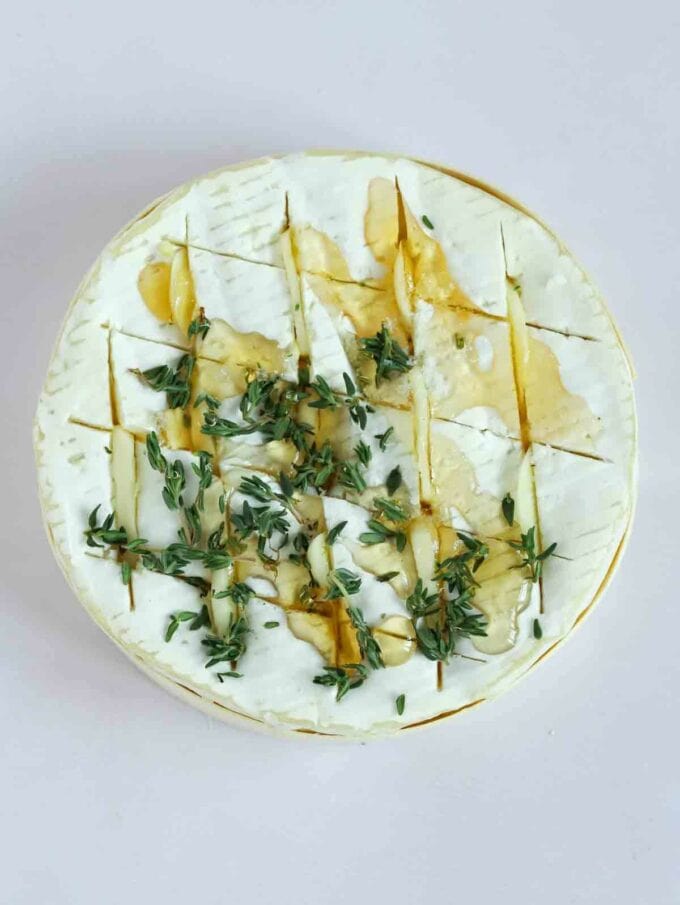 An uncooked camembert wheel with scored lines, with garlic slices, honey drizzled over and fresh thyme leaves sprinkled on top.