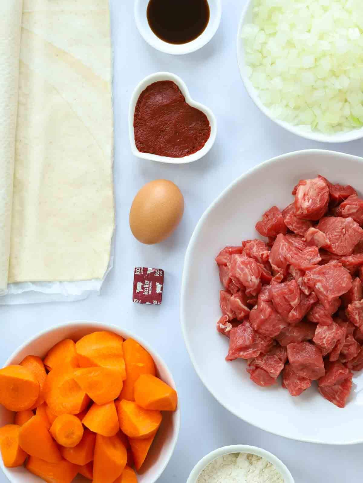 The ingredients for a steak pie recipe laid out on a white tablecloth, including diced beef, carrots, onion, egg, pastry, stock cube, tomato puree and Worcester sauce.
