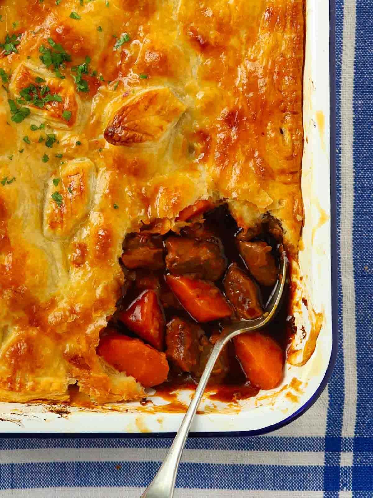 A blue and white tea towel with a pie dish on top, with a golden cooked steak pie being served up with a spoon.