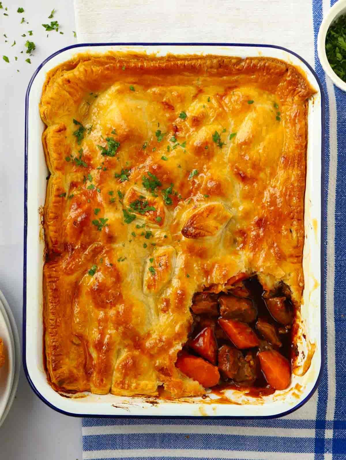 A steak pie in a dish with a golden crust with a piece missing, showing the beef and carrot filling. The pie sits on top a table with a blue and white tea towel under it.