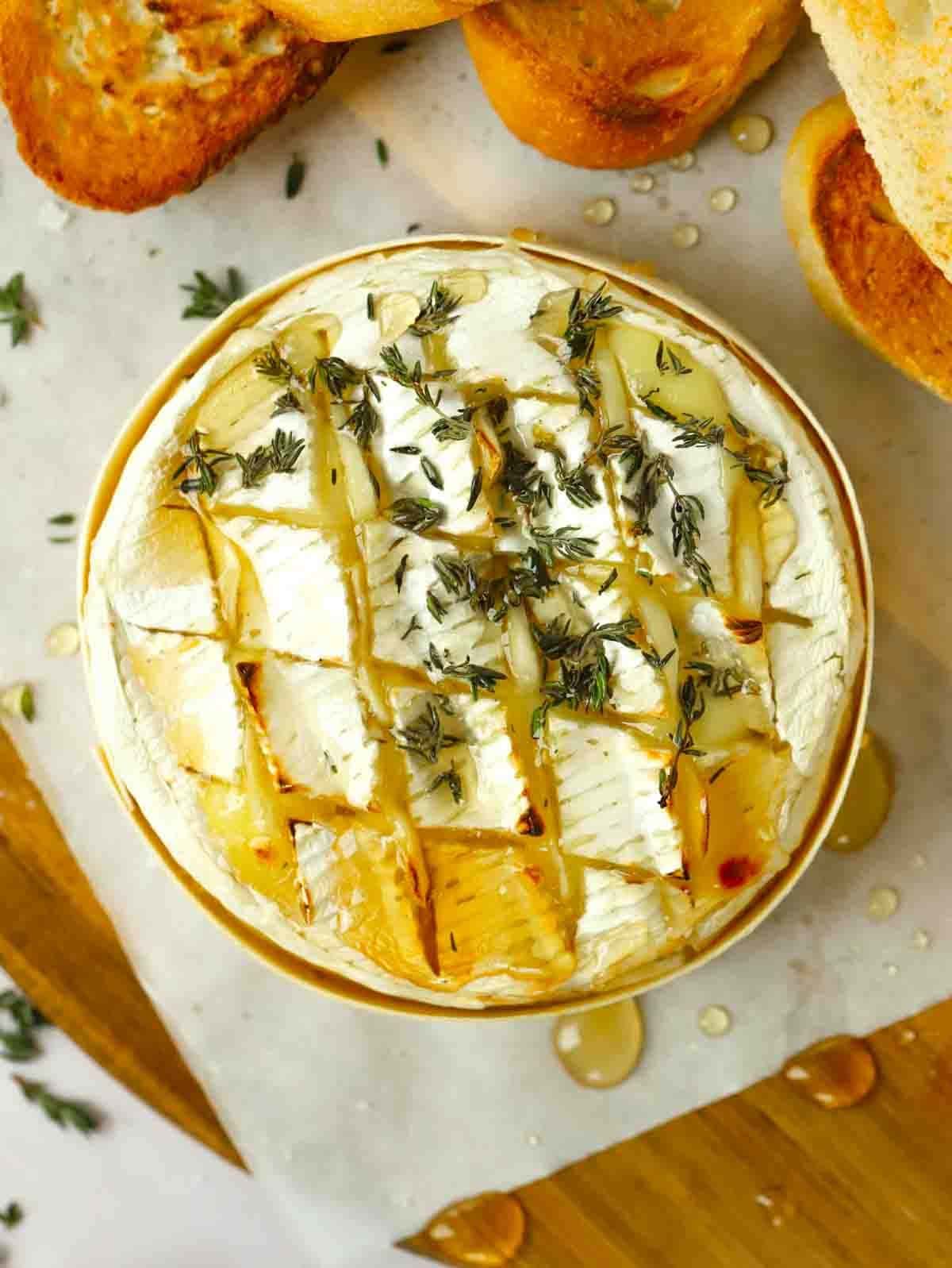 A baked round of camembert inside a wooden case, on a table with toasted bread at the side. Honey and thyme drizzled on top.
