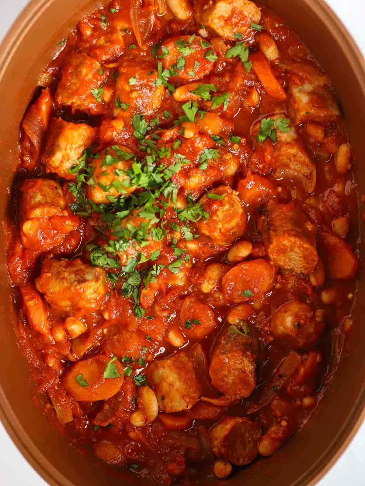 A slow cooker pan with a cooked sausage casserole inside, garnished with parsley.