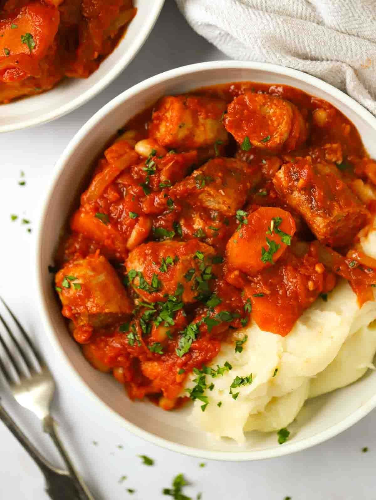 A bowl of sausage casserole and mashed potato, laid on a white table with a fork at the side.