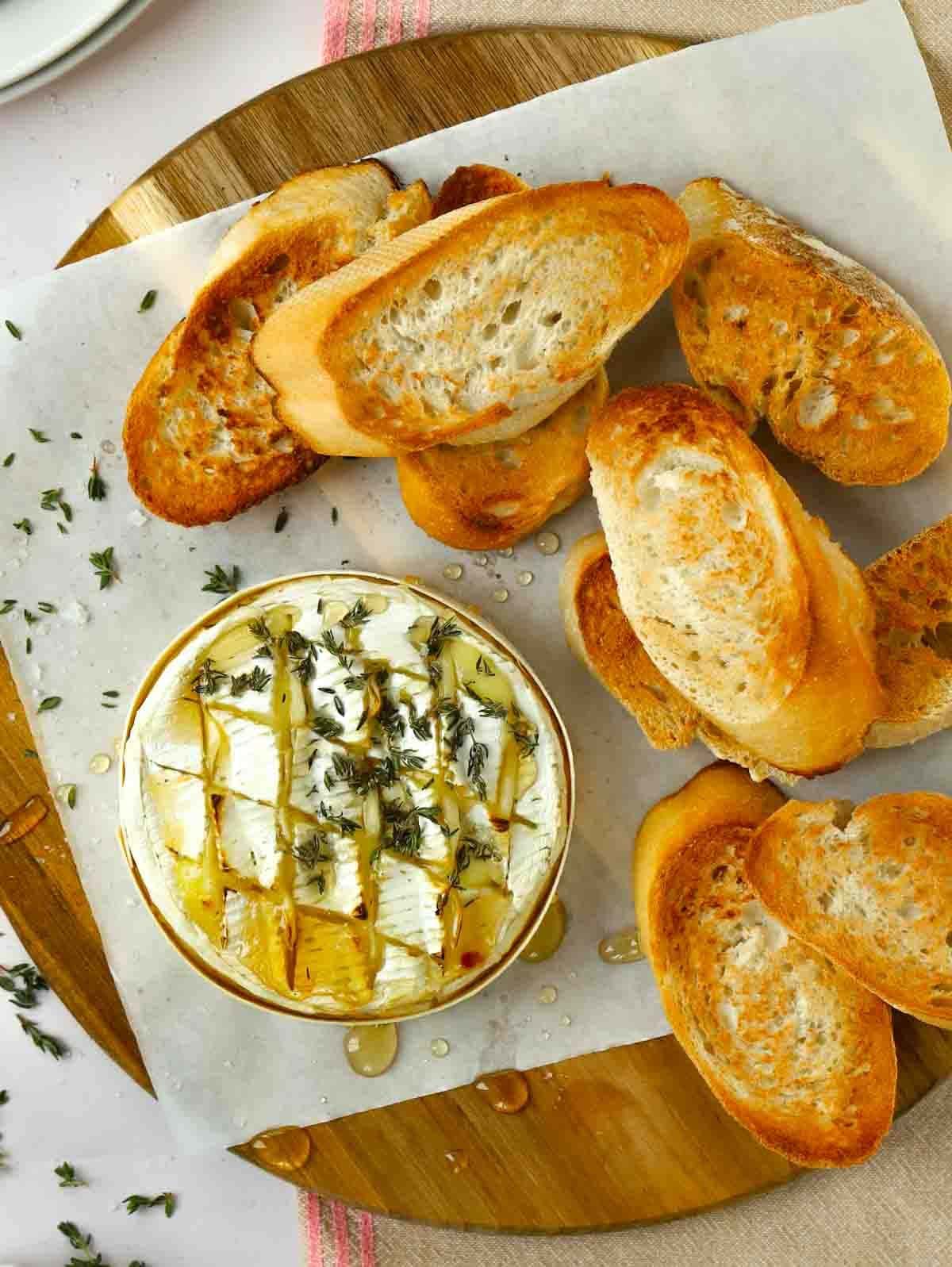 A round wooden board with sliced, toasted French bread on top with a round of baked camembert cheese.