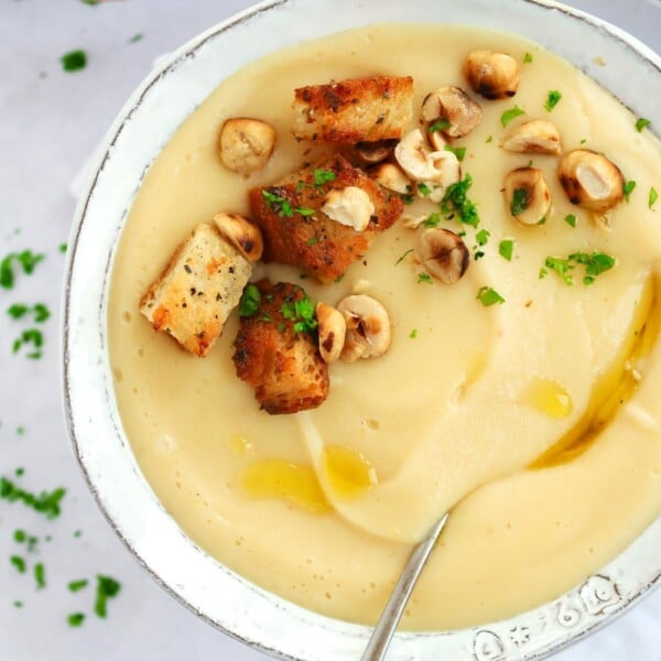 A bowl filled with creamy parsnip soup, with a spoon in and garlic croutons and halzenuts sprinkled on top. The bowl is on top of a white tablecloth.