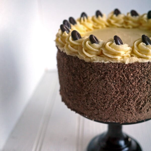 A Tiramisu Cake on a cake stand, with chocolate icing and a buttercream frosting on top, decorated with chocolate coffee beans.