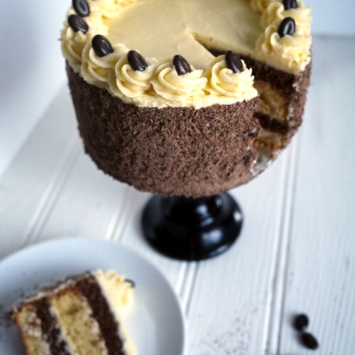 A layered Tiramisu Cake on a cake stand on a white surface with a portion missing, with a slice of cake on a plate at the side.