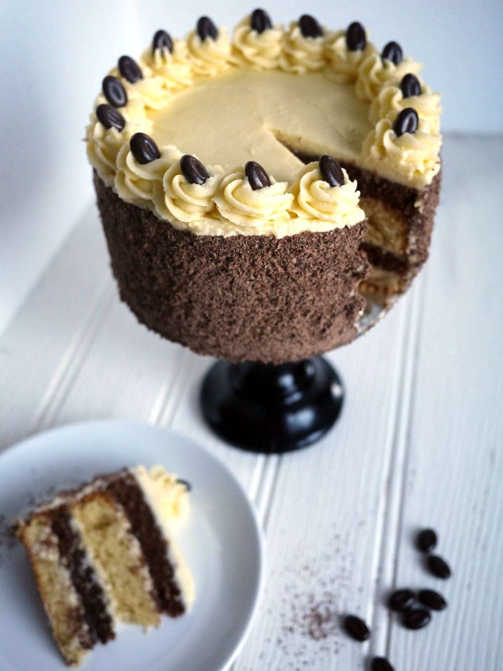 A layered Tiramisu Cake on a cake stand on a white surface with a portion missing, with a slice of cake on a plate at the side.