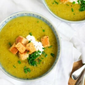 A bowl of Courgette Soup with toppings of chives, creme fraiche and croutons, with another bowl slightly in shot, both on a white tablecloth.