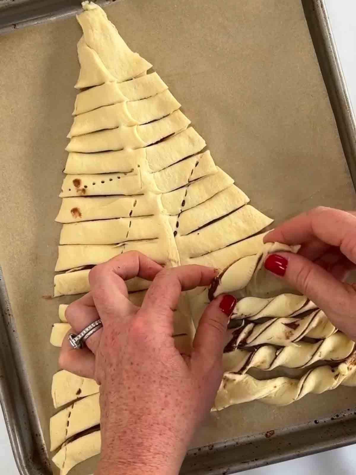 A triangle of croissant dough with strips in the sides, with two hands twisting the strips. For the recipe Nutella Christmas Tree.