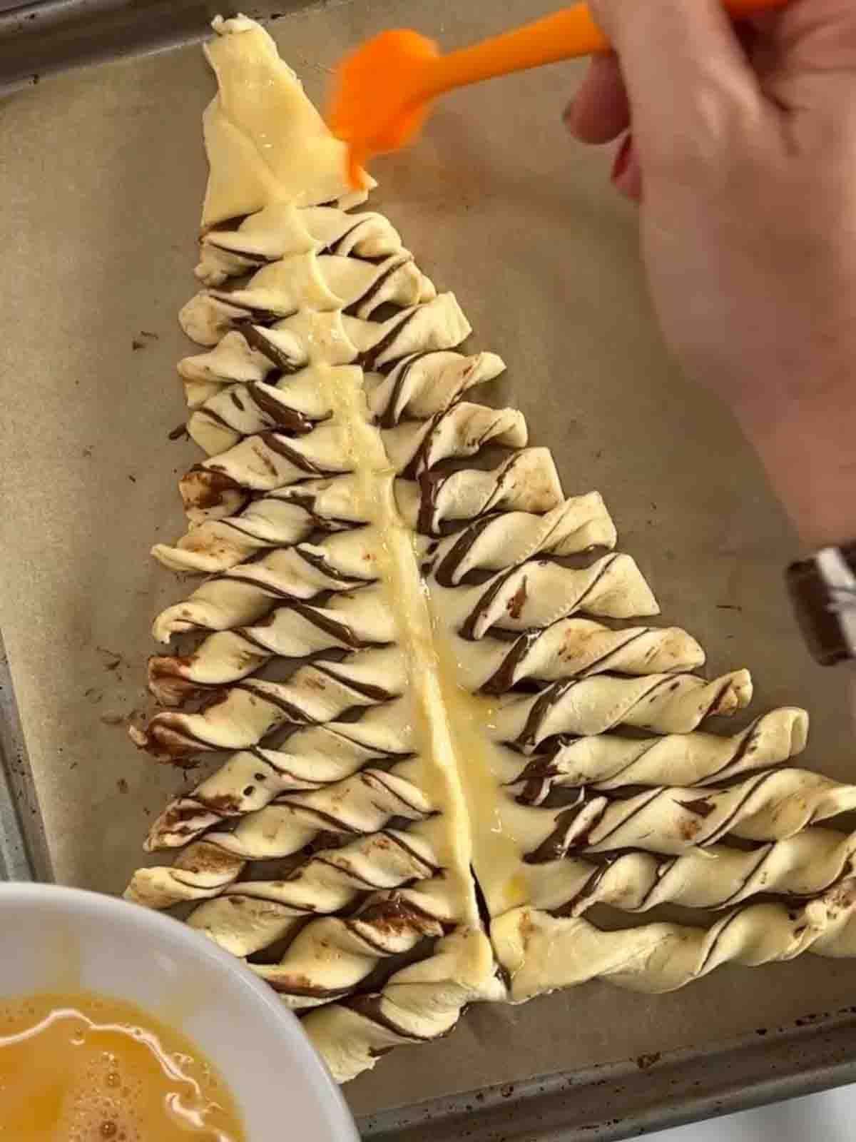 An unbaked Nutella Christmas Tree on a baking tray being brushed with egg before it goes in the oven.